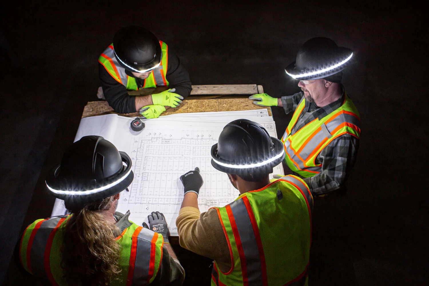 Four workers wearing Halo lights gather around plans on a jobsite