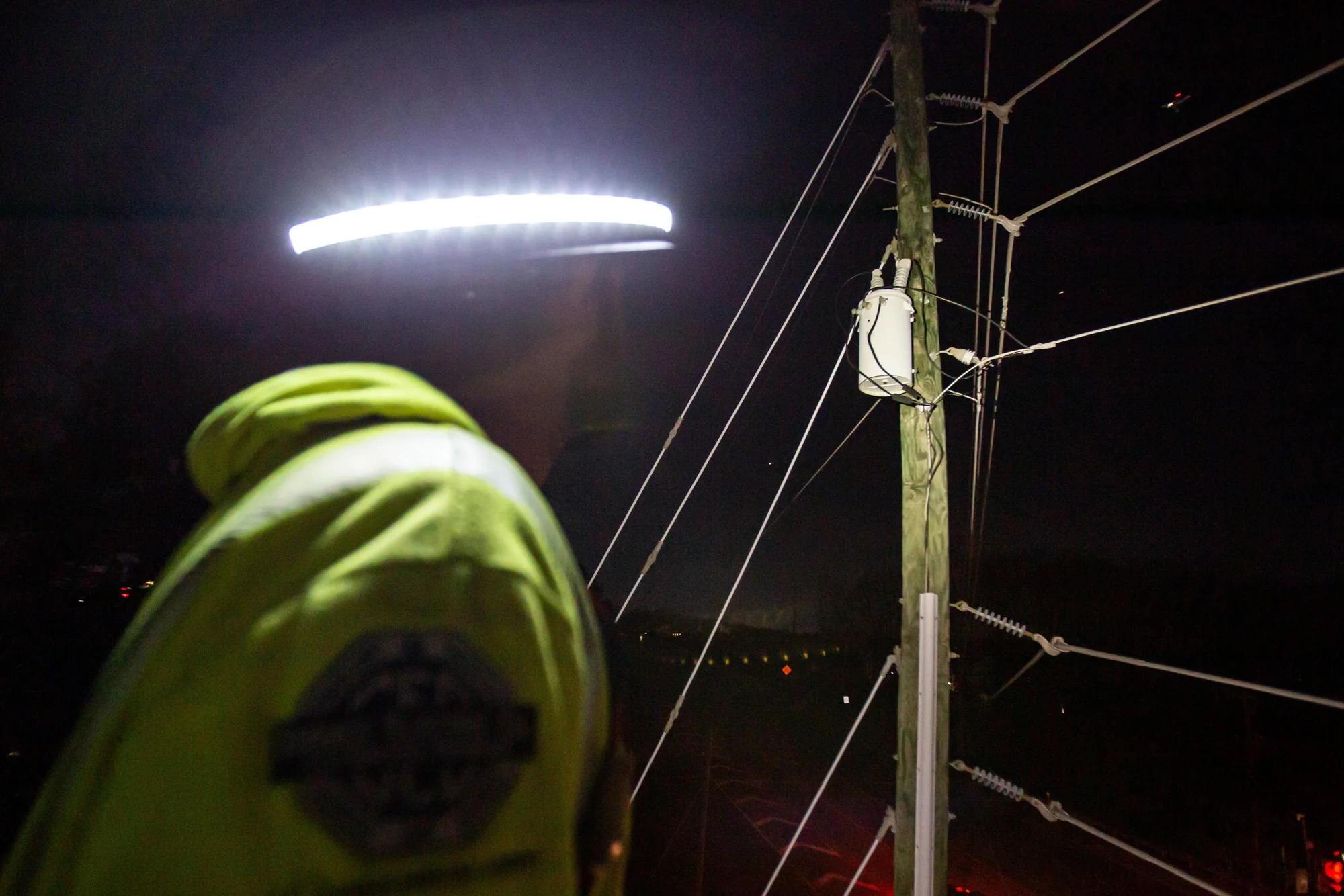 A utility worker wearing an illuminated Halo on a hard hat beside power lines at night